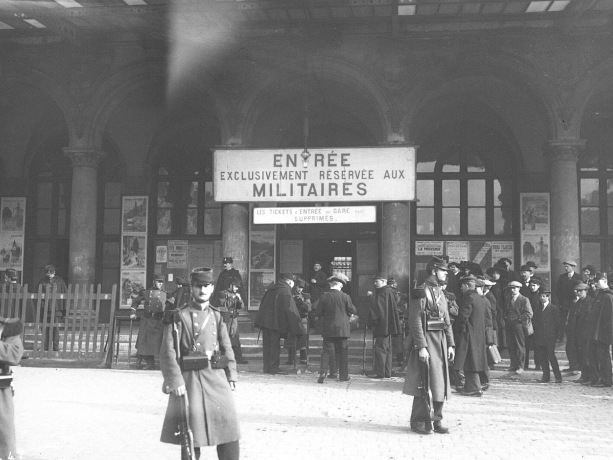 Devant une gare parisienne à arcades, deux soldats en capote montent la garde pendant qu'une foule d'hommes en manteaux attend sous un panneau d'entrée