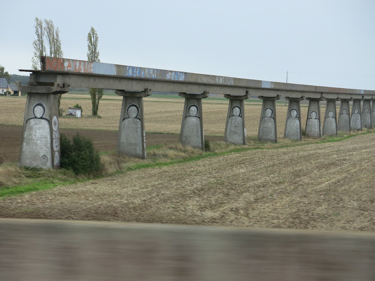 Rangée de piles de béton marquées de silhouettes peintes, portant une poutre de monorail qui s'interrompt net au-dessus d'un champ