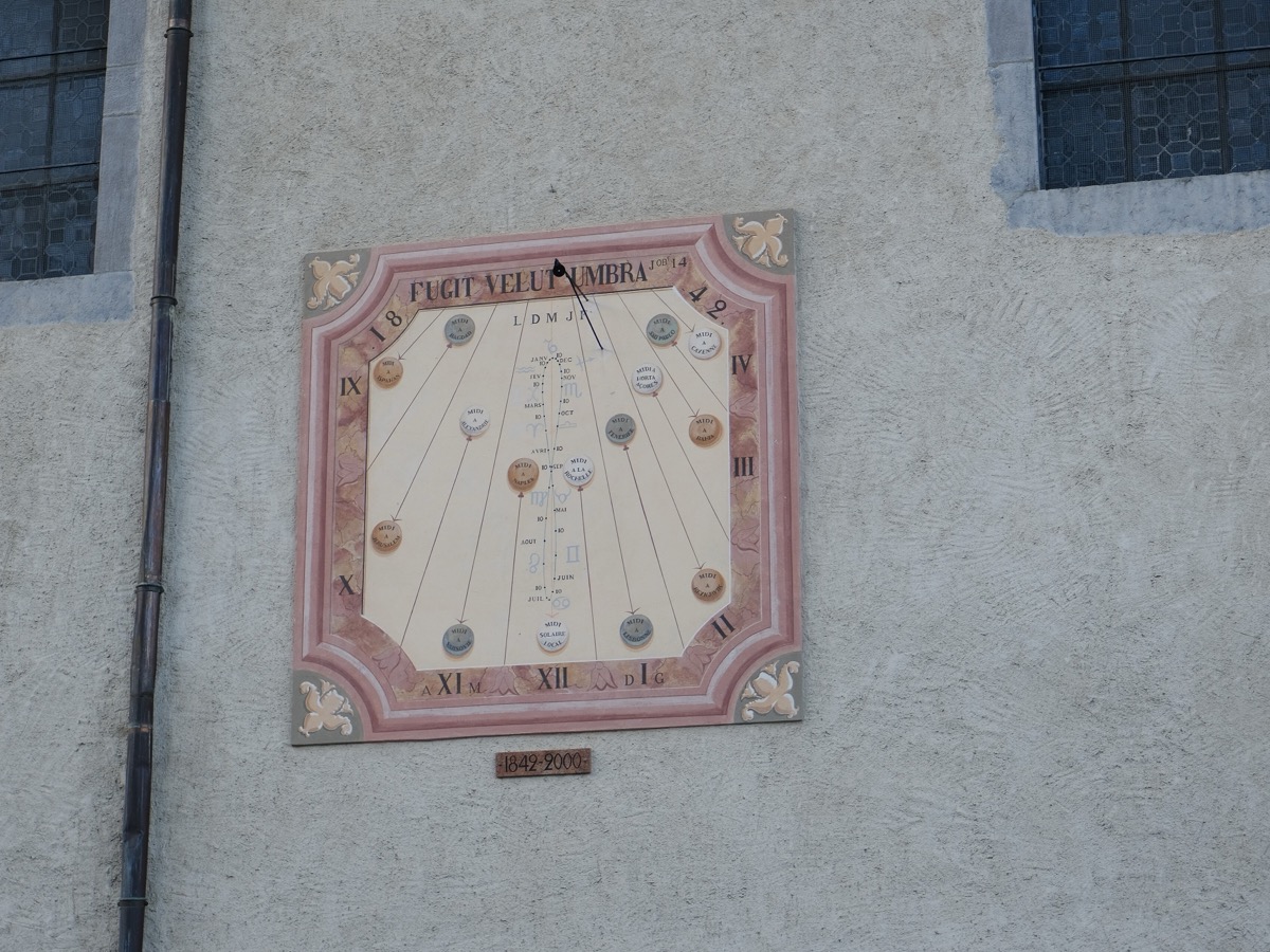 Cadran solaire peint sur le mur clair d'une église, à décor rose et fleurs de lys, avec ses chiffres romains et l'ombre du style au milieu de la journée