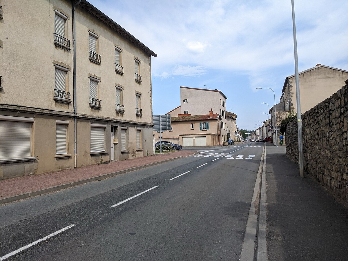 Route nationale traversant un bourg, immeubles anciens à volets roulants sur la gauche, passage piéton et lampadaires, chaussée vide en légère montée