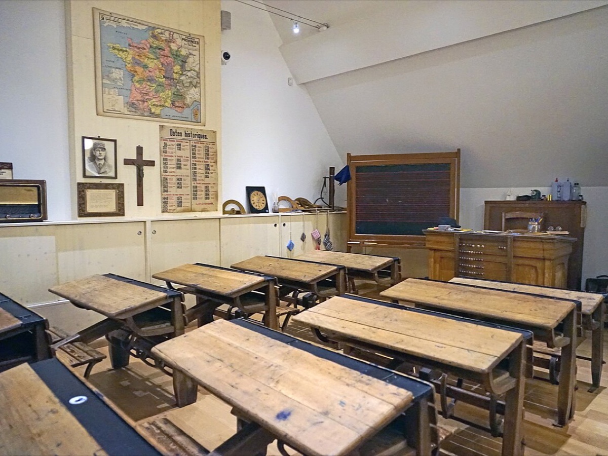 Salle de classe reconstituée avec des pupitres en bois à bancs solidaires, un tableau noir sur estrade, une carte de France murale et une affiche de dates historiques