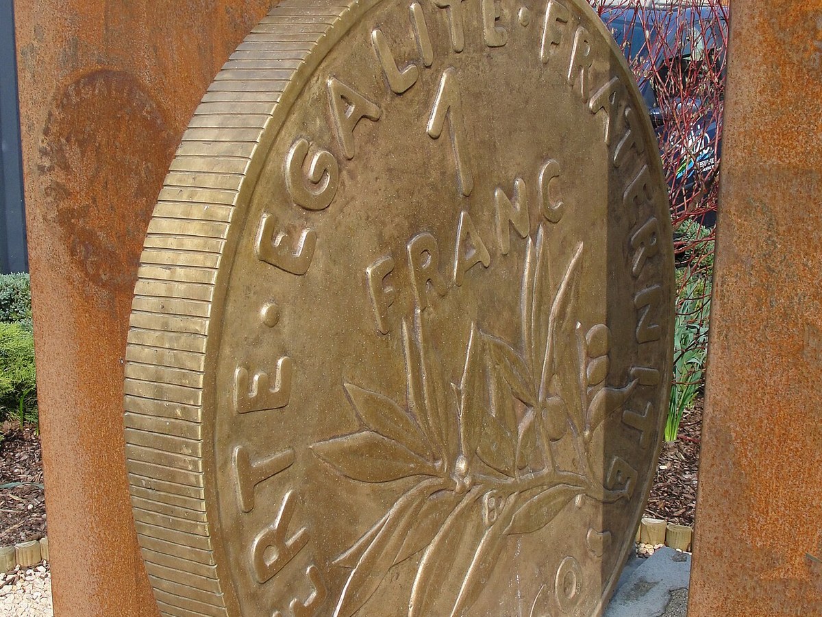 Reproduction monumentale en bronze d'une pièce de 1 franc, dressée entre deux montants d'acier rouillé dans un jardin de village