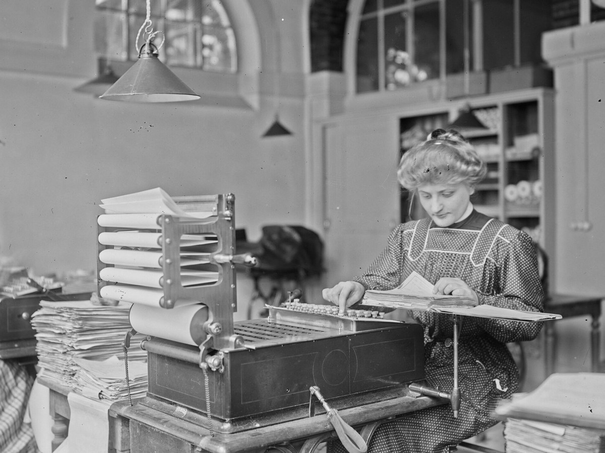 Une employée assise devant une machine mécanique à cartes perforées, entourée de piles de formulaires, dans un bureau du début du vingtième siècle