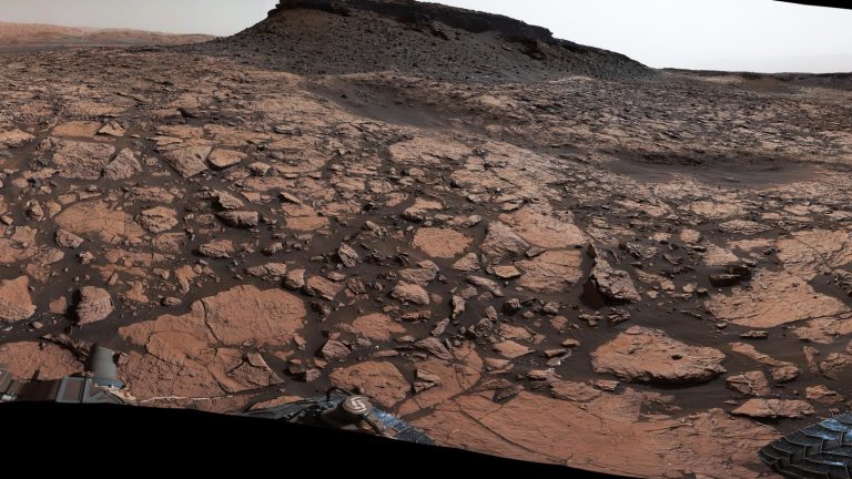 Paysage martien de dalles rocheuses et de buttes sombres sous un ciel clair, vu depuis la surface