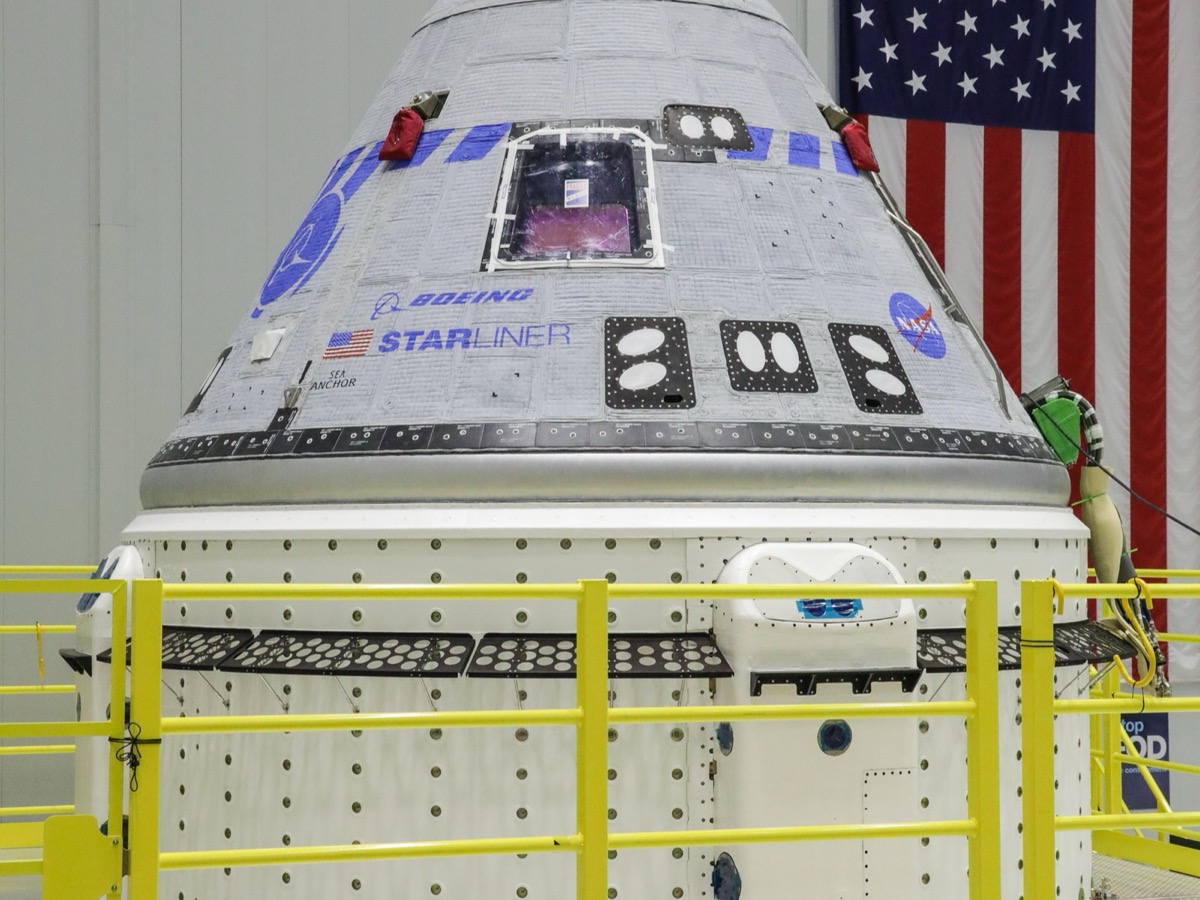 Capsule Starliner posée sur son module de service cylindrique blanc dans un hall d'assemblage, derrière une barrière jaune