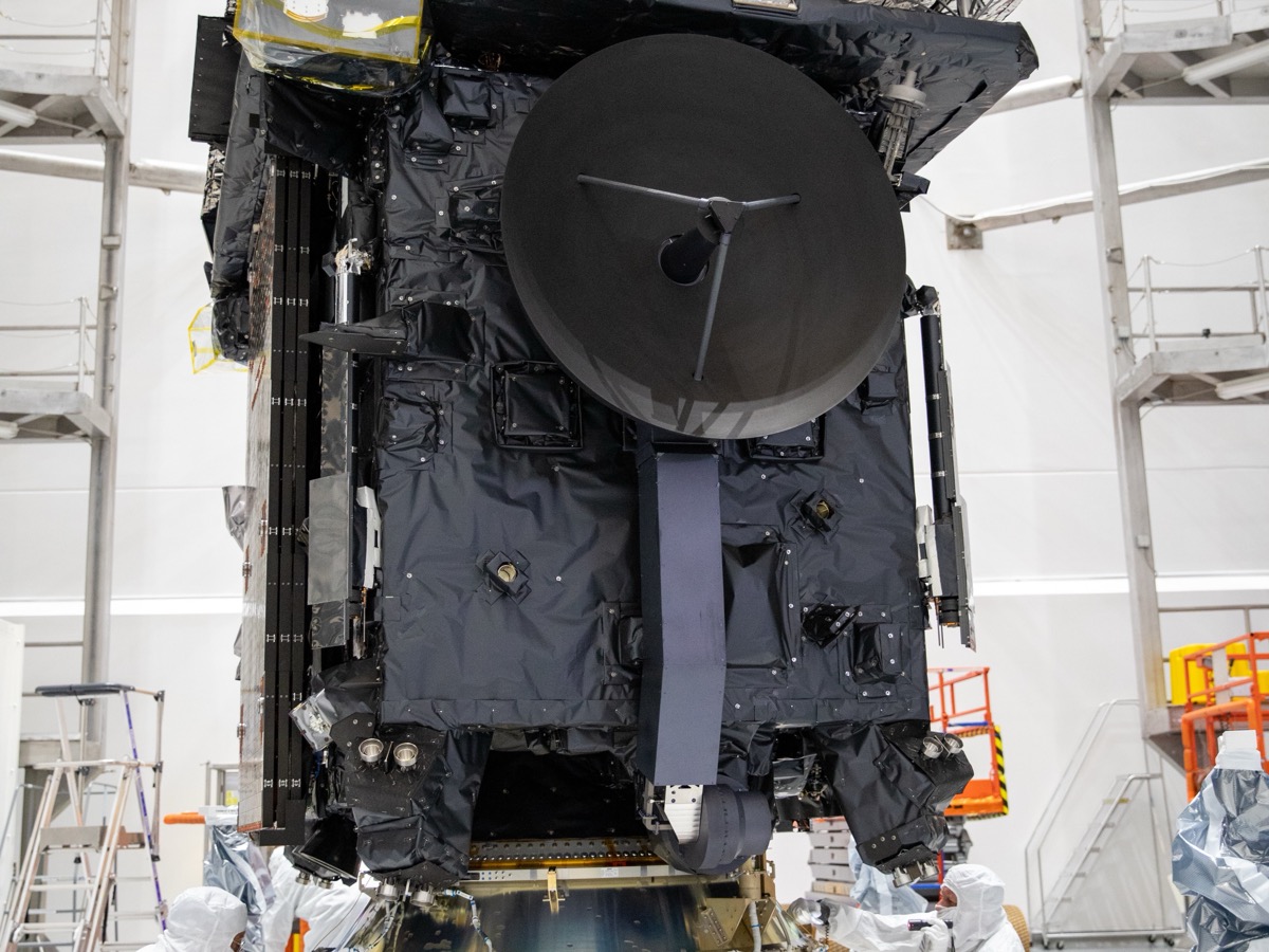 Sonde spatiale emballée dans ses couvertures noires, antenne ronde au premier plan, dans un hall d'assemblage