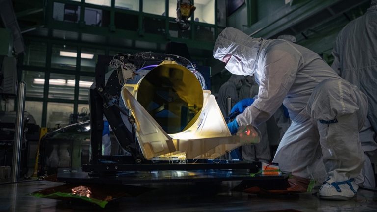Technicien en combinaison de salle blanche penché sur un télescope dont le miroir doré est visible
