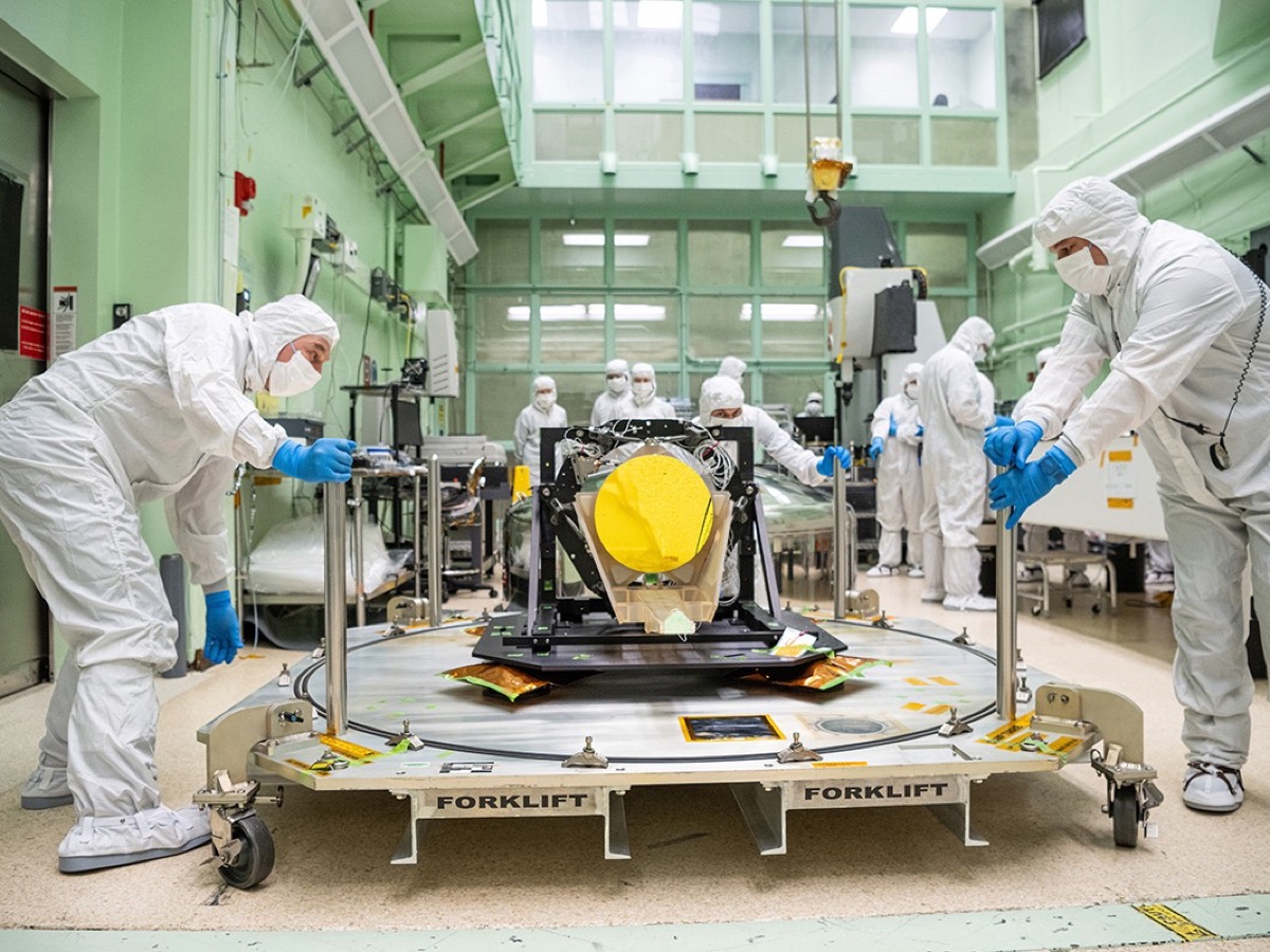 Techniciens en combinaison blanche déplaçant un télescope à miroir doré dans une salle blanche