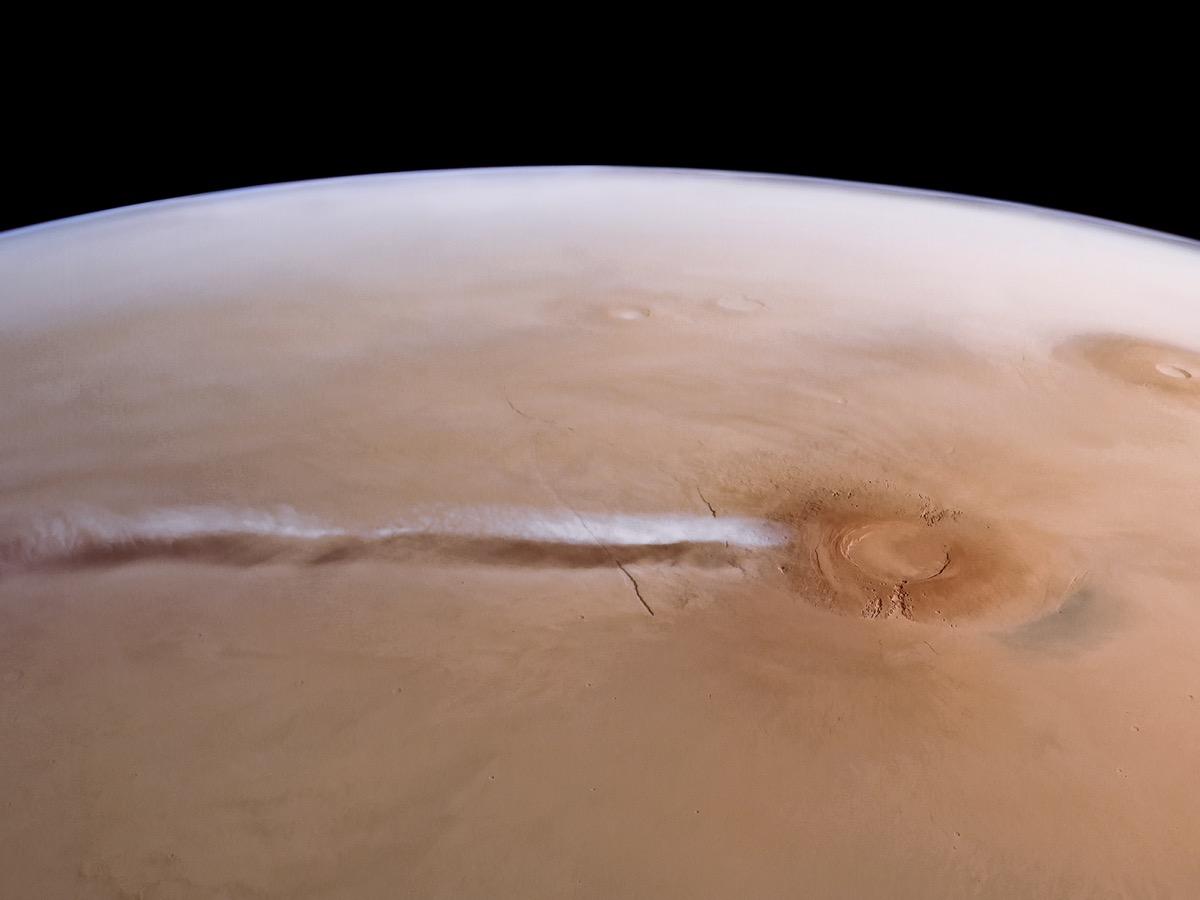 Vue oblique de la planète Mars montrant une longue traînée blanche étirée depuis un volcan, avec son ombre portée au sol