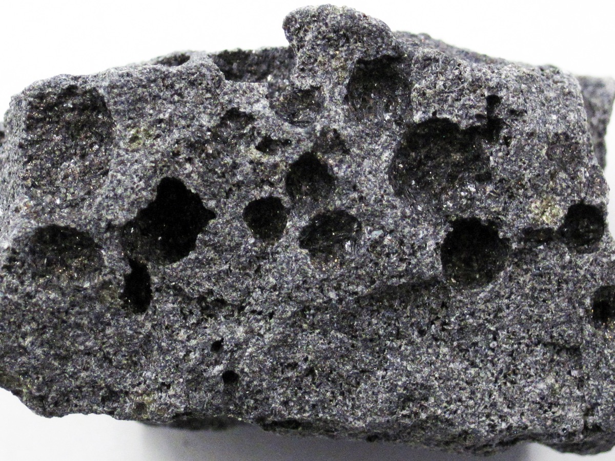 Morceau de basalte gris sombre criblé de cavités arrondies laissées par des bulles de gaz, posé sur fond blanc