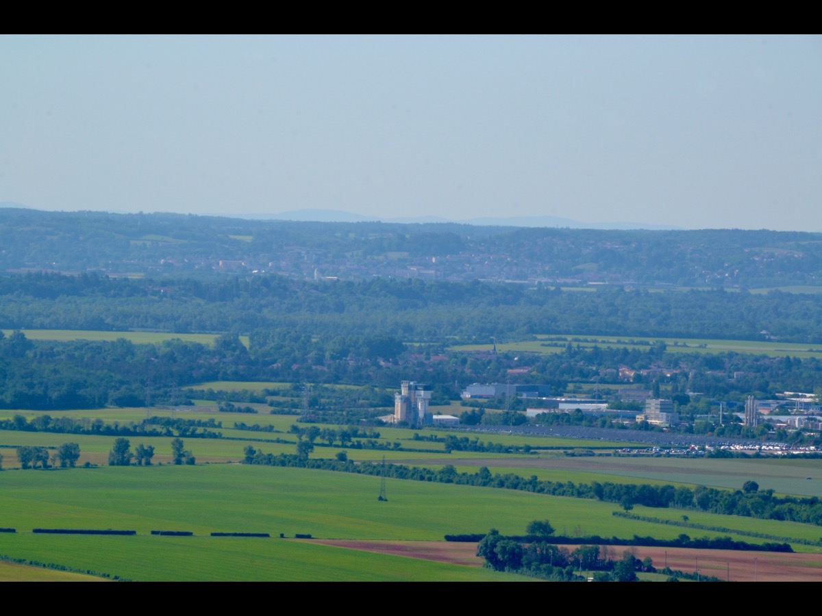 Vue d'une plaine avec des champs, des bâtiments industriels et des silos alignés au second plan, sous un ciel clair