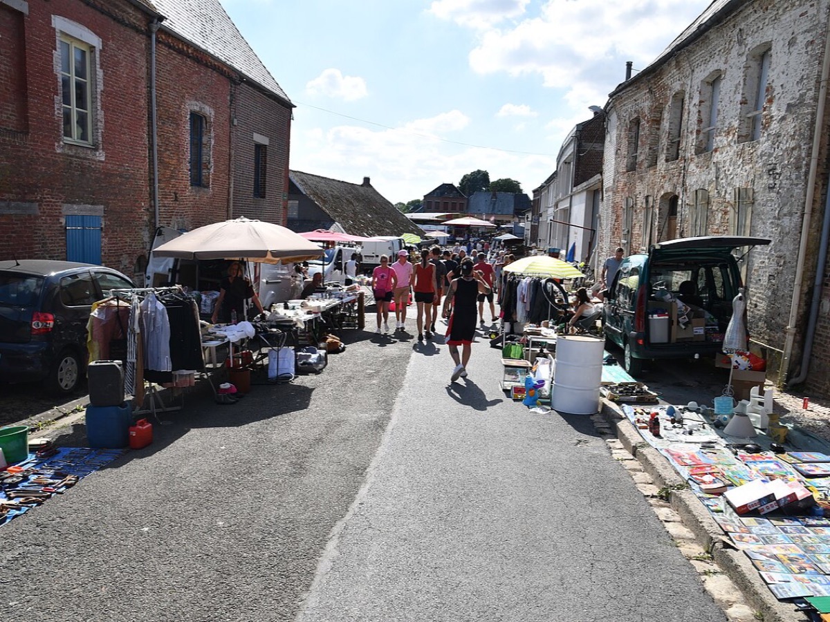 Rue de village bordée de stands de brocante, objets exposés sur des tables et à même le sol