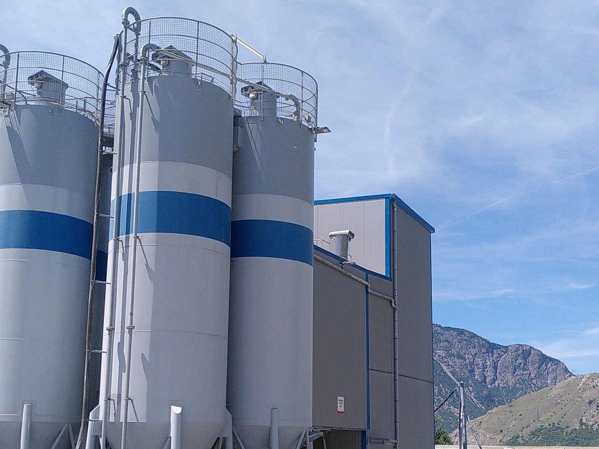 Quatre silos gris et bleus d'une centrale à béton adossés à un bâtiment métallique, montagne en arrière-plan
