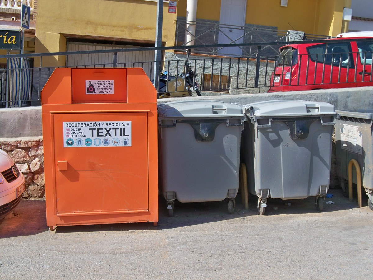 Conteneur de collecte textile orange installé à côté de deux bacs à ordures ménagères gris, dans une rue en pente