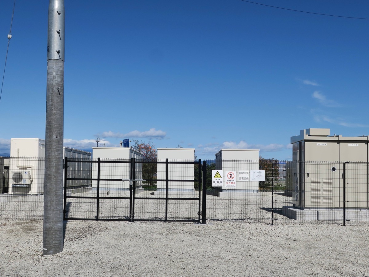 Rangée de conteneurs de stockage par batteries derrière une clôture grillagée, au bord d’une route de campagne