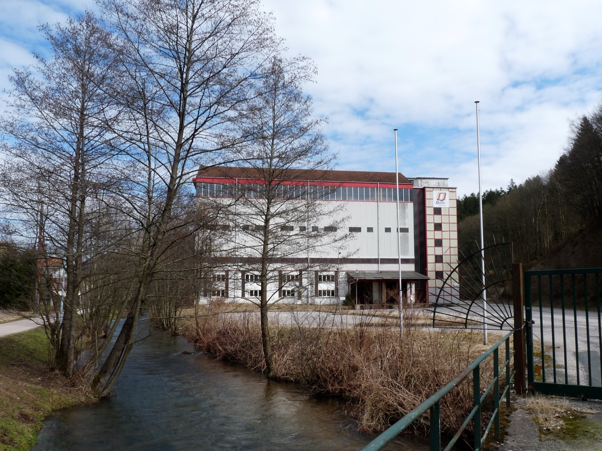 Bâtiment industriel blanc à bandeau rouge, au bord d’un ruisseau, dans une vallée boisée des Vosges
