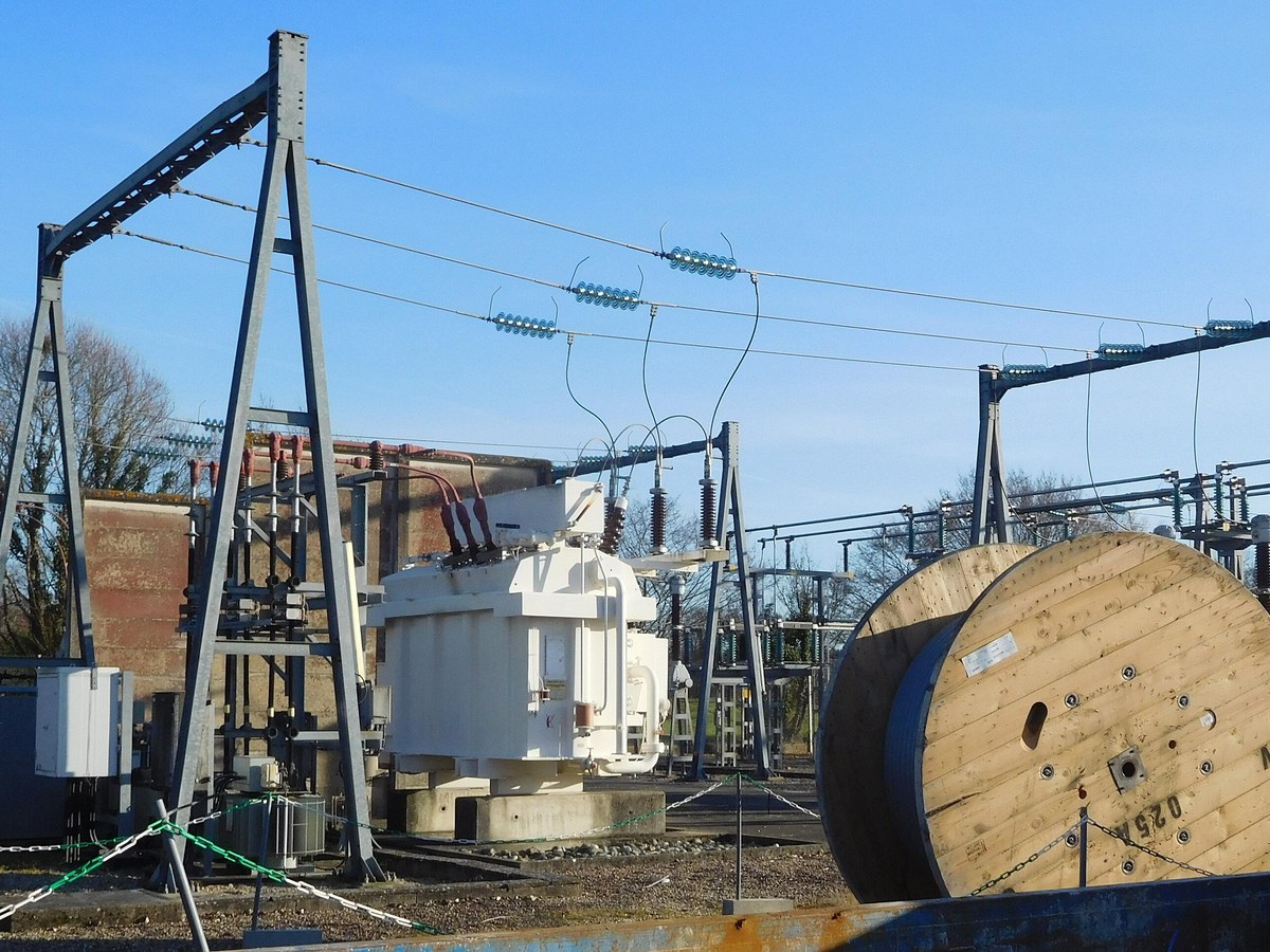 Poste électrique haute tension avec portiques métalliques, isolateurs et transformateur blanc, sous un ciel bleu