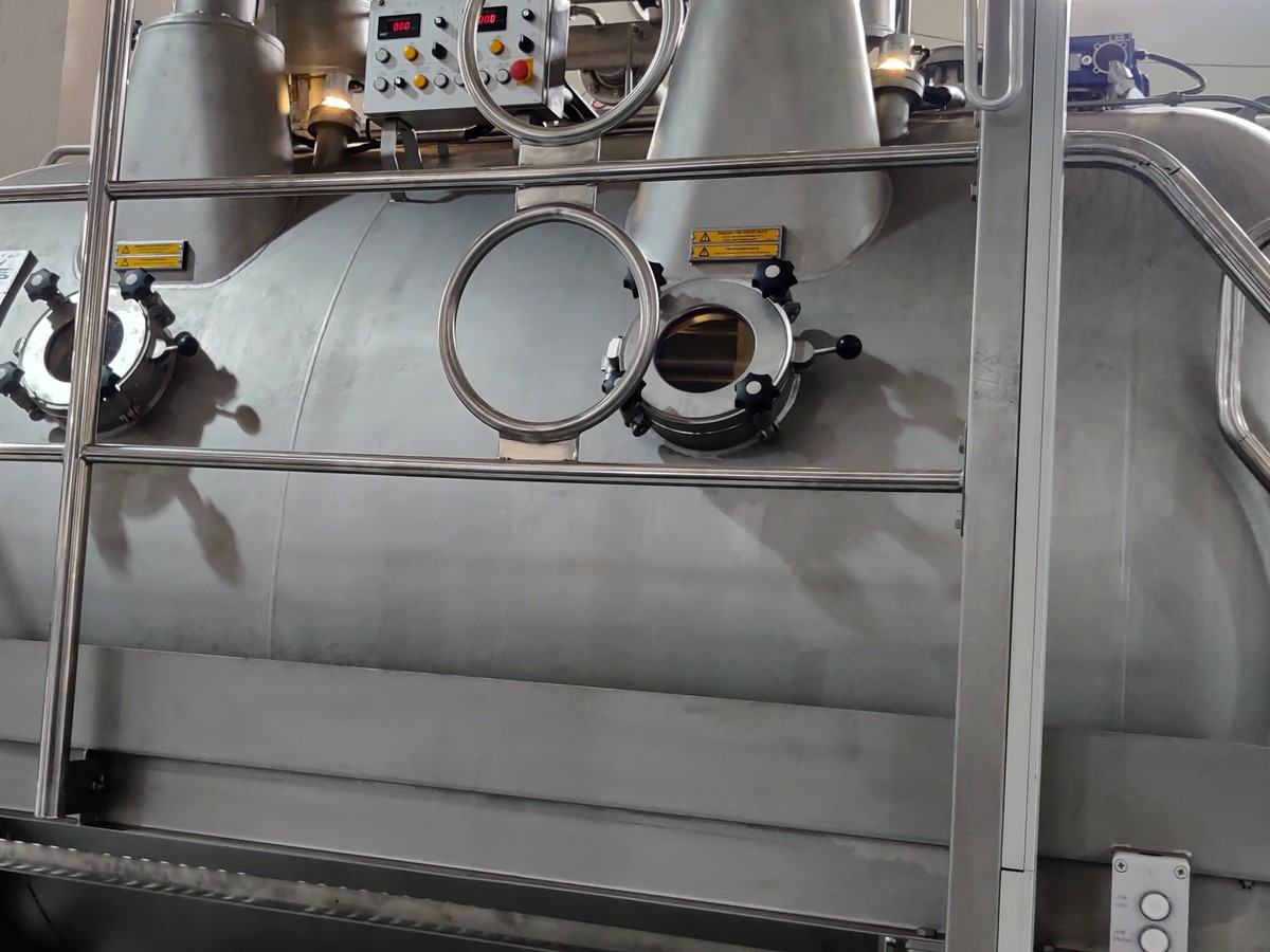 Grande machine cylindrique en inox dans un atelier textile, avec hublots de visite, passerelle et tableau de commande