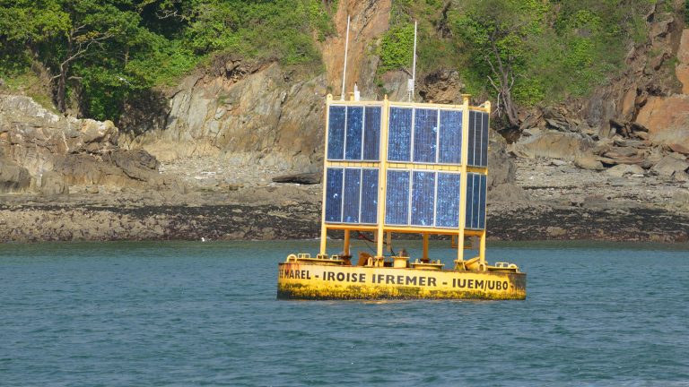 Bouée de mesure jaune portant deux rangées de panneaux solaires, au mouillage devant une côte rocheuse et boisée
