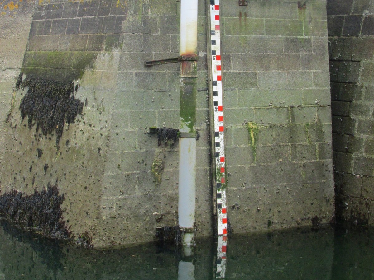 Échelle de marée graduée fixée contre un quai de pierre, à côté du tube blanc d'un marégraphe, au-dessus de l'eau