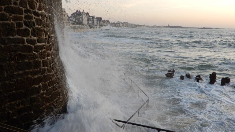 Une vague franchit le haut du rempart de Saint-Malo et retombe en nappe sur la promenade, la digue du Sillon en arrière-plan