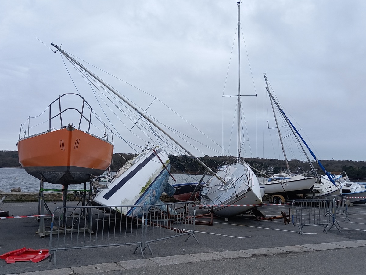 Voiliers couchés et appuyés les uns sur les autres sur le terre-plein d'un port, entourés de barrières de sécurité
