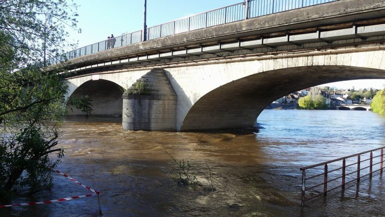 Rivière en crue aux eaux brunes passant sous un pont de pierre, une rambarde de berge à demi submergée
