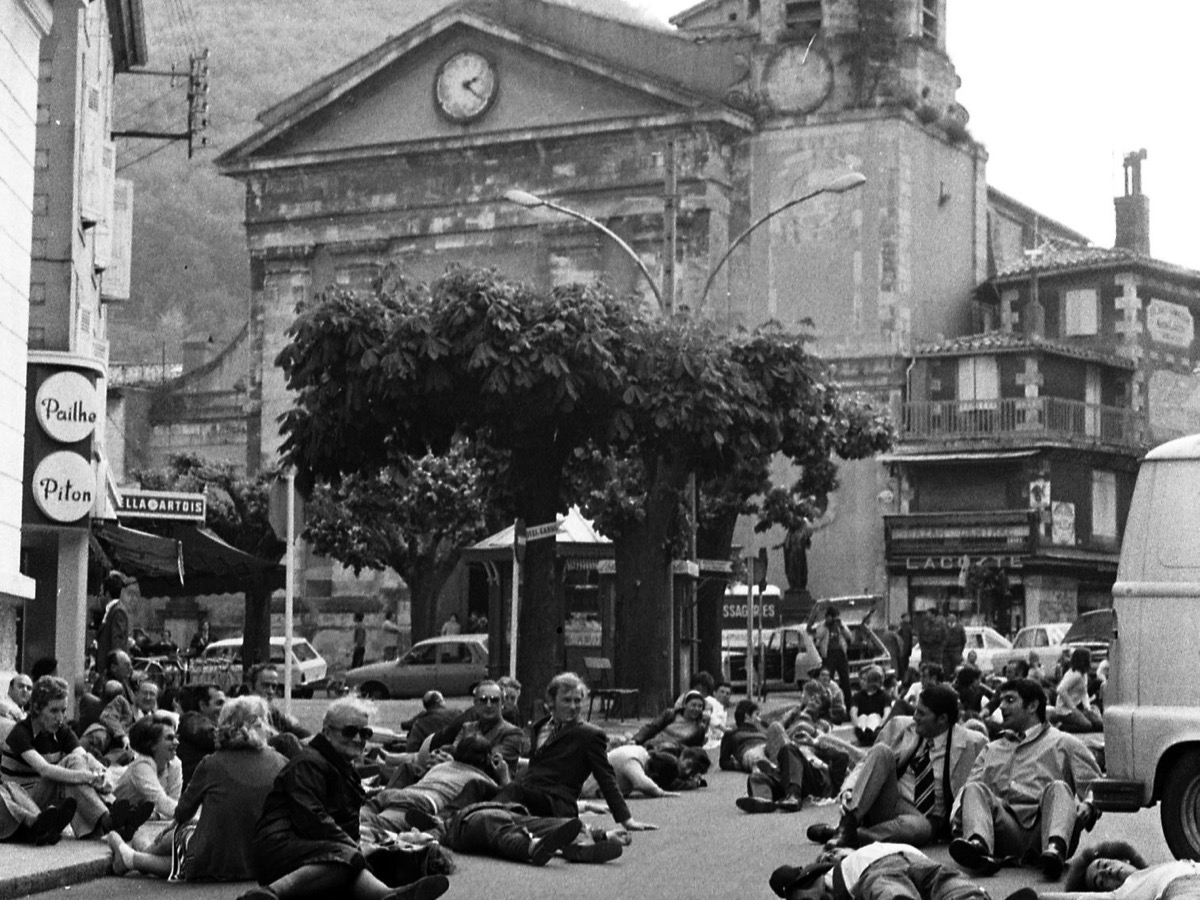Habitants allongés et assis sur la chaussée devant une église, voitures et commerces des années 1970 à l'arrière-plan