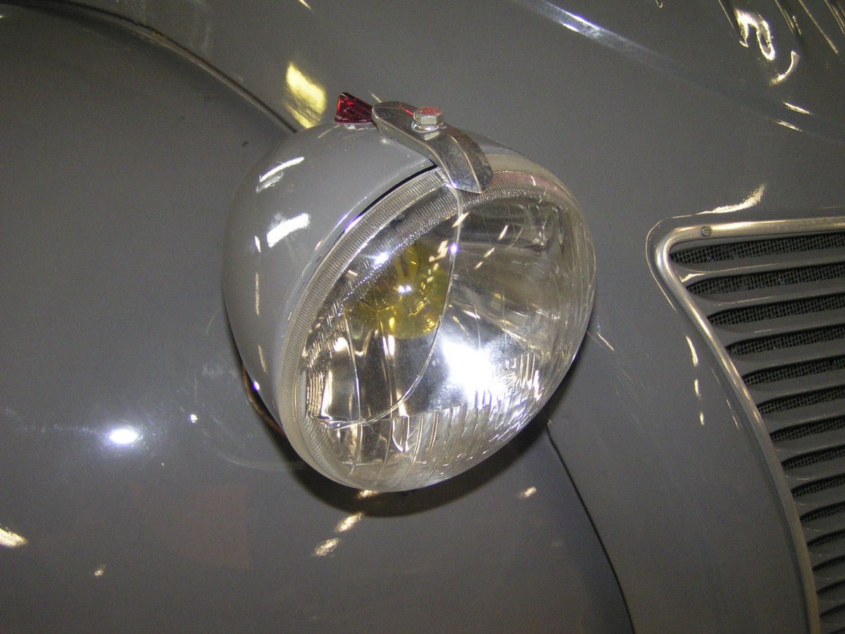 Gros plan sur le projecteur rond avant d'une Citroen 2CV grise, dont l'ampoule visible derriere la glace apparait jaune