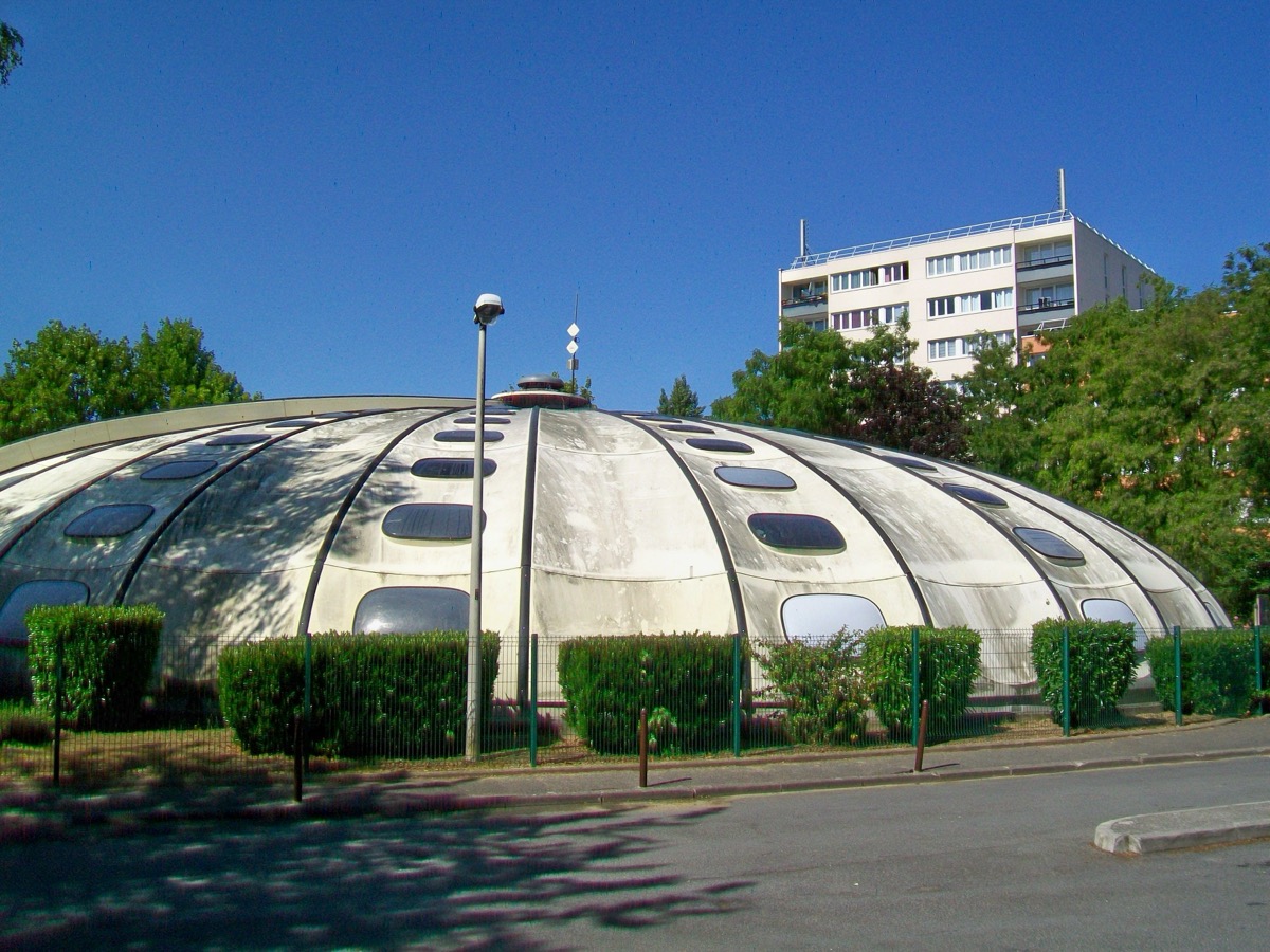 Piscine Tournesol de Fosses, coupole claire divisée en secteurs et percée de hublots ovales, derrière une haie et une grille