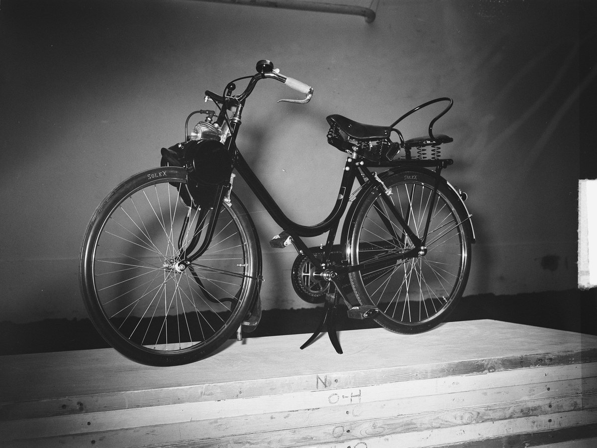 VéloSolex noir de 1951 posé sur une estrade de bois devant un mur clair, le moteur fixé au-dessus de la roue avant