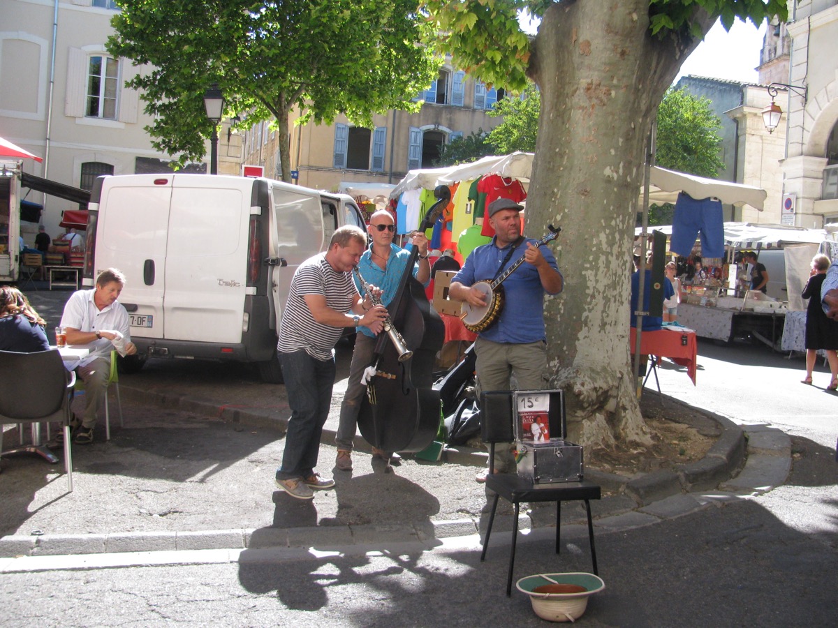Trois musiciens de rue jouent de la clarinette, de la contrebasse et du banjo sous un platane, au milieu d'un marché