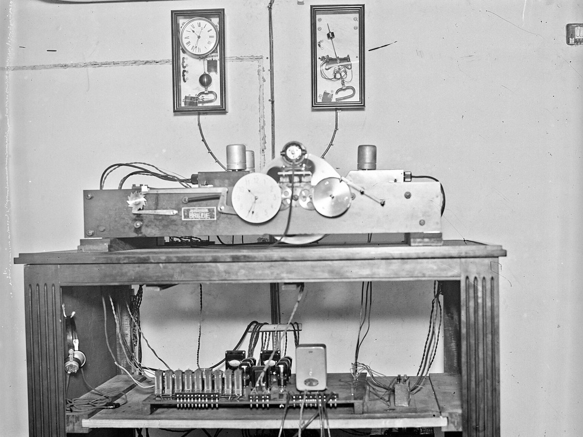 Installation de 1933 photographiée de face : une machine posée sur une table, des appareils électriques sur les étagères du dessous, deux pendules accrochées au mur
