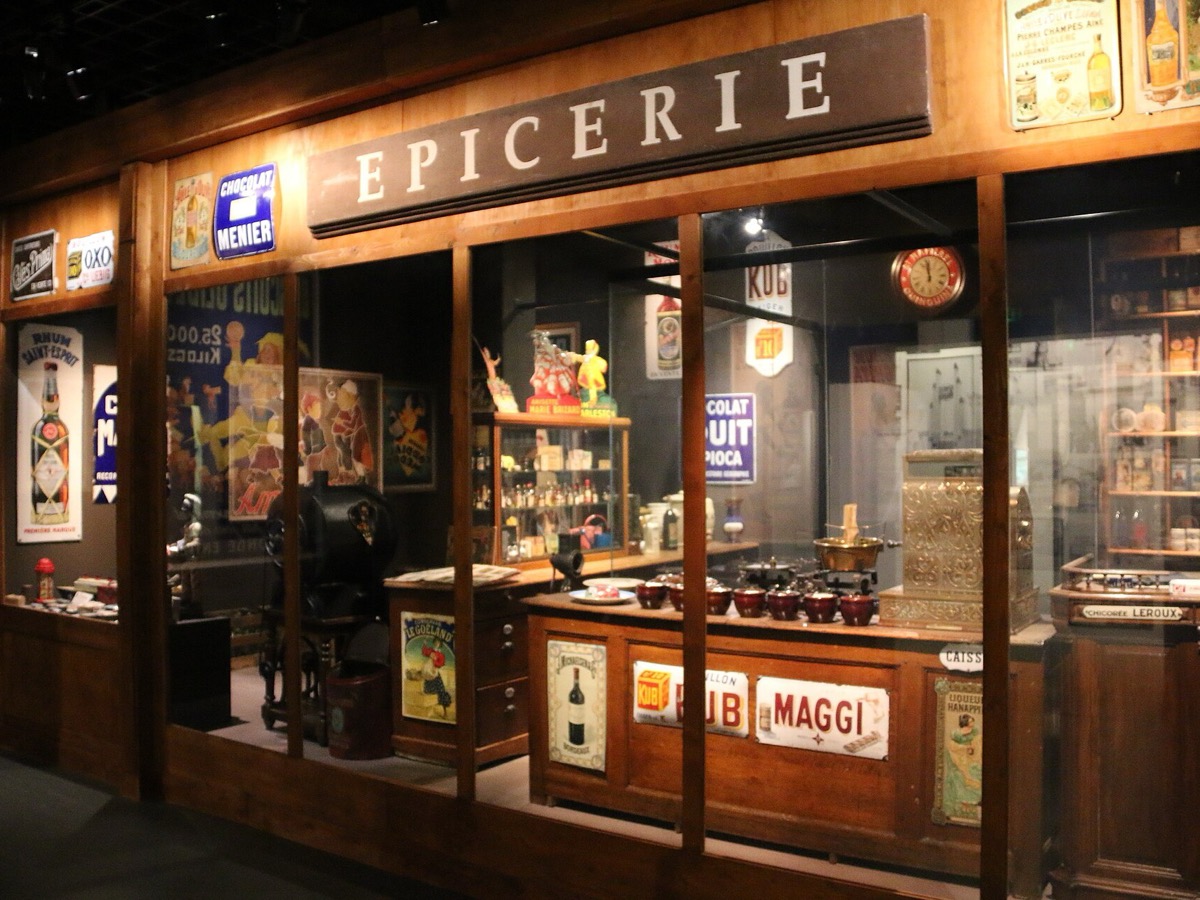 Devanture en bois reconstituee portant l'enseigne Epicerie, avec plaques emaillees, bocaux et caisse enregistreuse derriere une vitre de musee