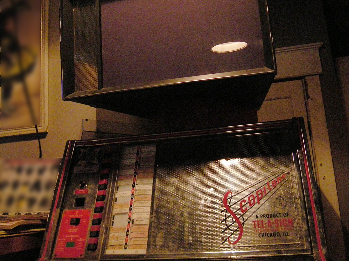 Juke-box à écran, avec un caisson de projection suspendu au-dessus d'un pupitre vitré garni de touches de sélection et d'étiquettes de titres