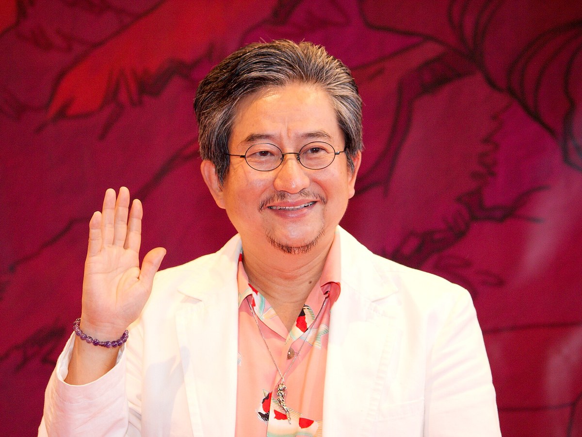 Portrait du mangaka Go Nagai, lunettes rondes et veste blanche, saluant de la main devant un fond rouge