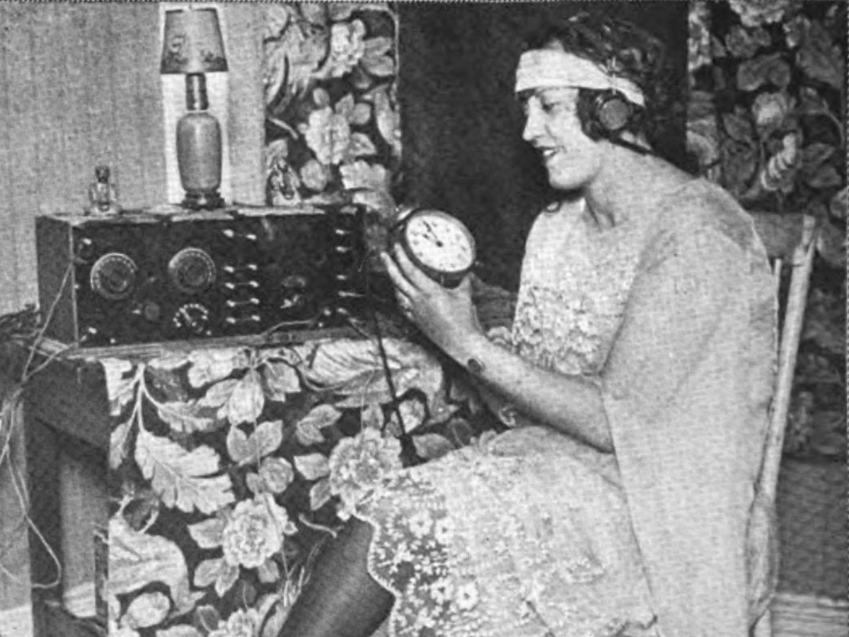 Une jeune femme coiffée d'un casque d'écoute tient un réveil devant un poste de radio, photo de 1922
