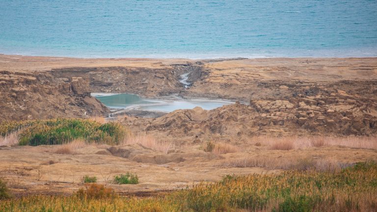 Doline effondrée remplie d'eau turquoise sur l'ancien rivage de la mer Morte, avec le lac en arrière-plan