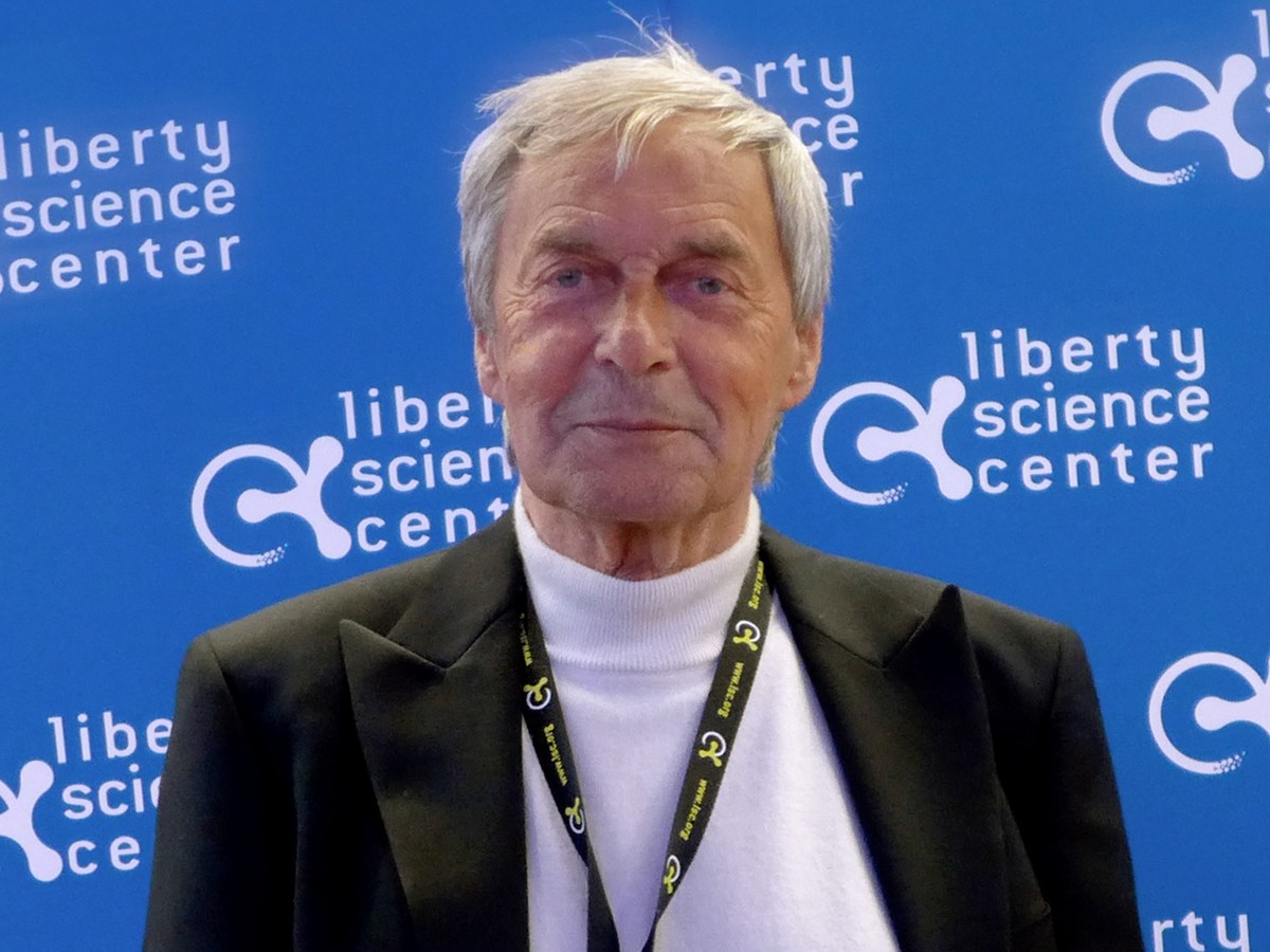 Portrait d'Ernő Rubik aux cheveux blancs, en veste sombre et col roulé blanc, devant un panneau bleu